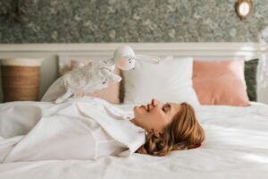 A cheerful woman lies on a bed, joyfully holding a plush puppet in a cozy bedroom setting.